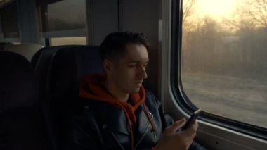 Tren yolculuğu sırasında Smartphone kullanan genç adam ve güzel gün batımı ile pencerenin karşısında oturuyor