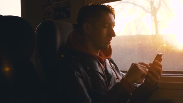 Fiatal férfi okostelefon segítségével vonattal lovagolni, és ül szemben az ablakon, gyönyörű naplemente — Stock videók