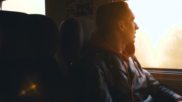 Молодой человек смотрит у окна на закате, а затем на камеру во время путешествия на поезде — стоковое видео