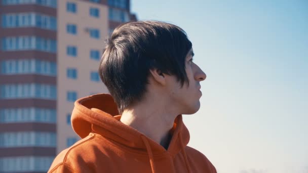 Retrato de un joven con una sudadera con capucha roja en el fondo de un edificio de varios pisos — Vídeos de Stock