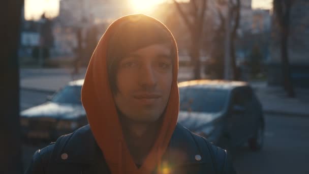 Närbild av stilig kaukasiska man med långt hår som bär casual kläder tittar på kamera vid solnedgången Urban bakgrund. — Stockvideo
