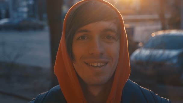 Retrato de homem caucasiano bonito feliz com capuz vermelho sorrindo na cidade ao pôr do sol urbano fundo — Vídeo de Stock