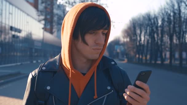 Joven guapo en la soleada calle urbana de la ciudad. Tipo con capucha roja usando teléfono inteligente, fondo de la puesta del sol . — Vídeo de stock