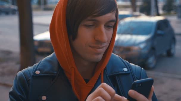英俊的年轻人在阳光明媚的城市街道城市背景。男子在红色连帽衫使用智能手机，日落背景. — 图库视频影像