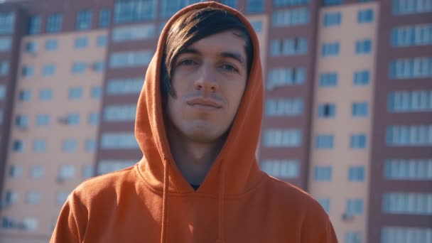 Portréja egy fiatal férfi egy piros kapucnis pulcsit a háttérben egy többszintes épület — Stock videók