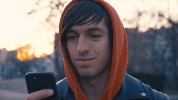 Joven guapo en la soleada calle urbana de la ciudad. Tipo con capucha roja usando teléfono inteligente, fondo de la puesta del sol . — Vídeos de Stock