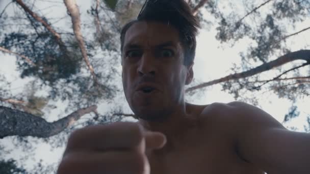 Безумный агрессивный мужчина без рубашки бьет кулаками лежащего в лесу человека — стоковое видео