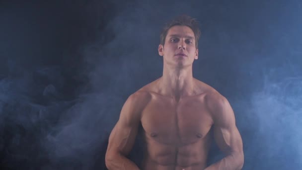 Hombre musculoso sin camisa posando sobre fondo oscuro, con humo a su alrededor . — Vídeos de Stock