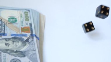 Yavaş çekimde iki siyah zar, boktan, beyaz arka plana atılmış, bir dolar paketinin yanında. Kumar kavramı.