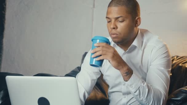 El hombre de negocios afroamericano usa computadora portátil y bebe café mientras está sentado en el sofá en la oficina moderna mira la pantalla del monitor — Vídeos de Stock