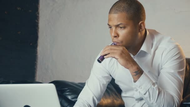 Σίγουροι καλά ντυμένος Αφρικανός Αμερικανός επιχειρηματίας χρησιμοποιώντας laptop και vaping ένα ηλεκτρονικό τσιγάρο στο γραφείο του, ενώ κάθονται στον καναπέ — Αρχείο Βίντεο