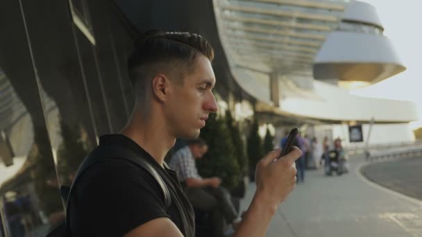 Attraente giovane uomo in t-shirt nera con il suo smartphone, nastro e scorrimento. All'aeroporto o alla stazione degli autobus . — Video Stock