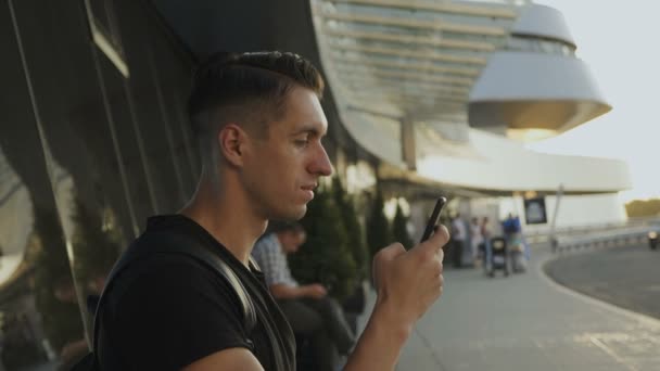 Homem jovem atraente usando seu smartphone, gravando e rolando, em seguida, virar a cabeça para a câmera e para trás. No aeroporto ou outro grande edifício de vidro — Vídeo de Stock