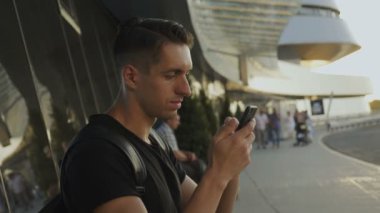 Siyah t-shirt çekici genç adam onun akıllı telefon kullanarak, kayıt ve kaydırma. Havaalanında veya otobüs durağında.