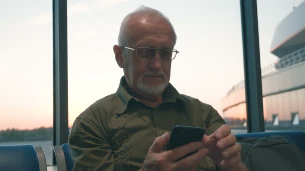 空港での搭乗を待つ灰色の髪と灰色のひげを持つシニア男性、スマートフォンを使用して時間を過ごす — ストック動画