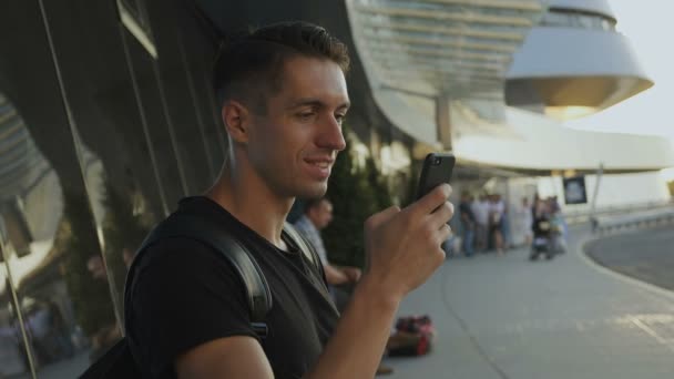 Ein lächelnder junger Mann in schwarzem T-Shirt bedient sein Smartphone, klebt und scrollt. am Flughafen oder Busbahnhof. — Stockvideo