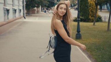 Çekici uzun saçlı woung kadın şehir-sokakta yürürken geri görünümü, kameraya döner ve güzel bir gülümseme verir, rüzgar saçları ile oynar. Steadicam atış.