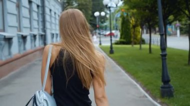 Çekici uzun saçlı woung kadın şehir-sokakta yürürken geri görünümü, kameraya döner ve güzel bir gülümseme verir, rüzgar saçları ile oynar. Steadicam atış.