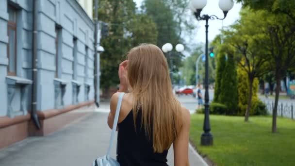 Back view of attractive long-haired woung woman walking down the city-street, turns to camera and gives a beautiful smile, wind plays with her hair. Steadicam shot. — Stock Video