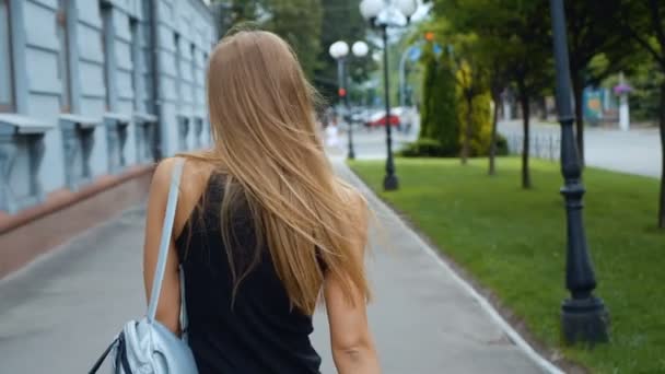 Vista posterior de la atractiva mujer de pelo largo caminando por la calle de la ciudad, se vuelve a la cámara y da una hermosa sonrisa, el viento juega con su cabello. Steadicam tiro . — Vídeos de Stock