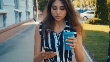 Şehir merkezinde yürürken akıllı telefon içeriğini kontrol ve kahve içme genç güzel kadın Ön görünümü.