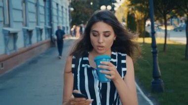 Şehir merkezinde yürürken akıllı telefon içeriğini kontrol ve kahve içme genç güzel kadın Ön görünümü.