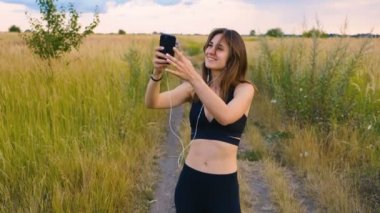 Mutlu genç kadın gün batımında sahada duran akıllı telefon ve kulaklık kullanın, sağlıklı yaşam tarzı. Akıllı telefon açık kullanarak Sportywoman koşucu.