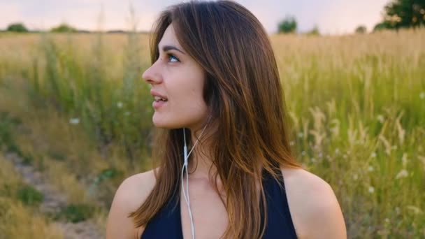 Mujer joven y feliz uso de teléfono inteligente y auriculares de pie en el campo al atardecer, estilo de vida saludable. Deportiva corredor utilizando el teléfono inteligente al aire libre . — Vídeos de Stock