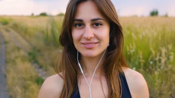 幸せな若い女性は、日没時にフィールドに立ってスマートフォンやヘッドフォンを使用し、健康的なライフスタイル。屋外のスマートフォンを使用してスポーティな女性ランナー. — ストック動画
