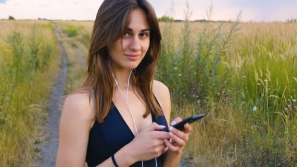 Lycklig ung kvinna använda smartphone och hörlurar stående i fält vid solnedgången, hälsosam livsstil. Sportywoman löpare använder smartphone utomhus. — Stockvideo