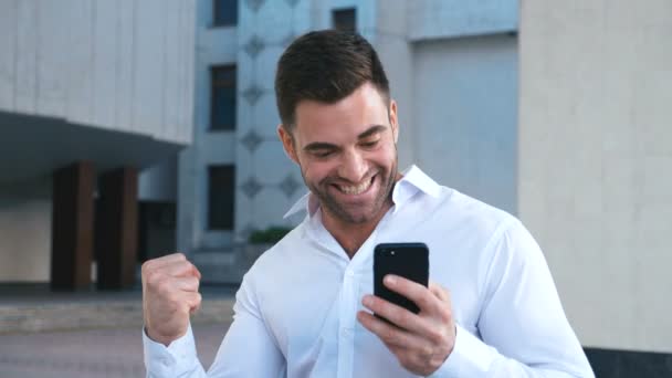Młody przystojny biznesmen obchodzi sukces podczas czytania wiadomości w smartphone w pobliżu budynku biurowego. Biznes, ludzie, komunikacja, technologia, Rozrywka i koncepcja stylu życia. — Wideo stockowe