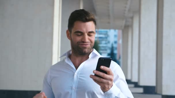 Empresario Celebrando el éxito mientras lee el mensaje en el teléfono inteligente cerca del edificio de oficinas. Guapo profesional exitoso hombre de negocios alcanzando objetivos personales . — Vídeo de stock