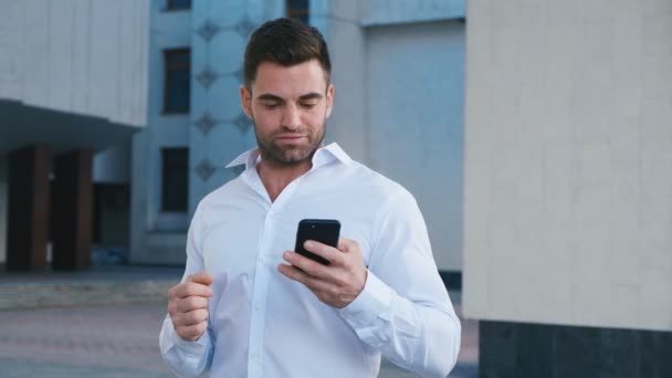 Obchodník oslavující úspěch při čtení zprávy v smartphone poblíž kancelářské budovy. Pohledný profesionální úspěšný obchodní muž, který dosáhl osobních cílů. — Stock video