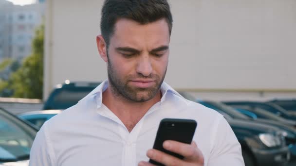 Joven hombre atractivo de pie en el estacionamiento cerca del edificio de oficinas escribiendo un mensaje en su teléfono inteligente. Hombre de negocios guapo en camisa blanca usando Smartphone al aire libre . — Vídeos de Stock