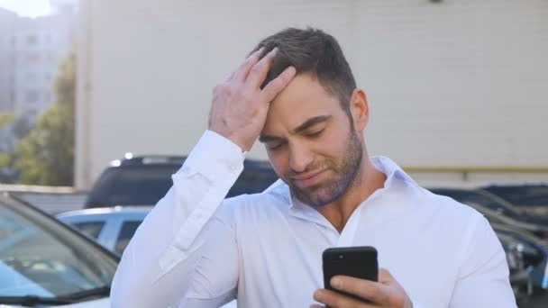 Joven hombre atractivo de pie en el estacionamiento cerca del edificio de oficinas escribiendo un mensaje en su teléfono inteligente. Hombre de negocios guapo en camisa blanca usando Smartphone al aire libre . — Vídeos de Stock