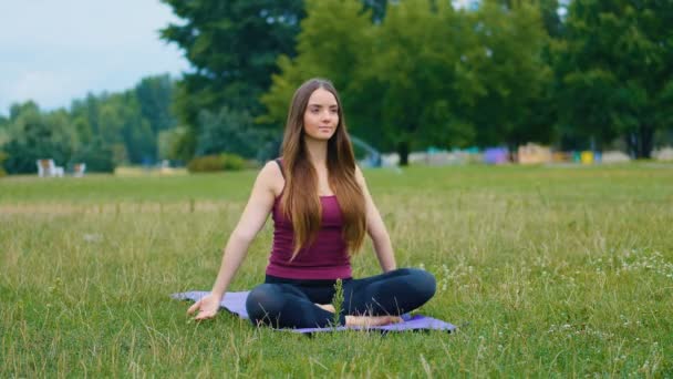 Mujer caucásica joven relajándose practicando yoga en el lugar al aire libre por la mañana. Hermosa mujer haciendo yoga en el parque en la hierba verde — Vídeo de stock