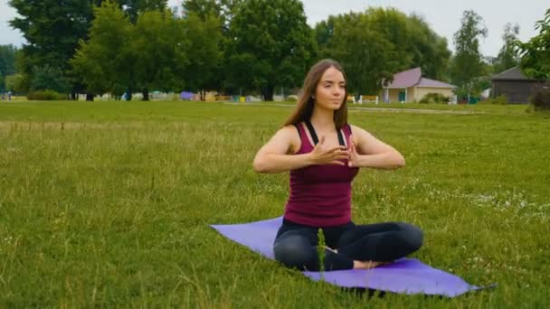 Mulher branca jovem relaxando praticando ioga no lugar ao ar livre na parte da manhã. Mulher bonita fazendo ioga no parque na grama verde — Vídeo de Stock
