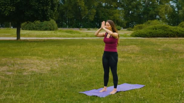 Νεαρή καυκάσια γυναίκα χαλαρώνει με την εξάσκηση γιόγκα σε υπαίθριο χώρο το πρωί. Όμορφη γυναίκα κάνει γιόγκα στο πάρκο στο πράσινο γρασίδι — Αρχείο Βίντεο