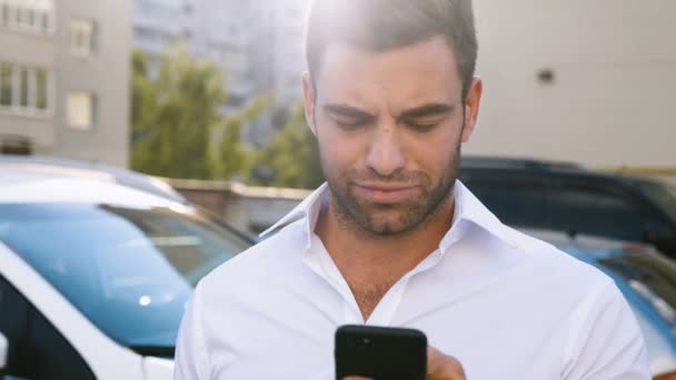 Ein junger attraktiver Mann steht auf einem Parkplatz in der Nähe eines Bürogebäudes und tippt eine Nachricht auf sein Smartphone. schöner Geschäftsmann im weißen Hemd mit Smartphone im Freien. — Stockvideo