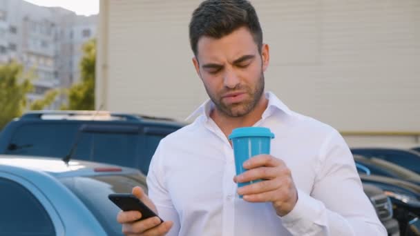 Stilig man använder smartphone och dricker läckra kaffe på en parkering i närheten av kontorsbyggnad. Ser nöjda. Affärs livsstil. — Stockvideo