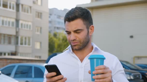 Bonito Homem Usando Smartphone e Beber Delicioso Coffe em um estacionamento perto de prédio de escritórios. Parece satisfeito. Estilo de vida do negócio . — Vídeo de Stock