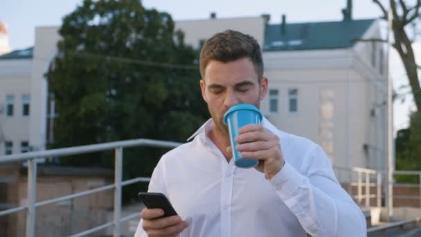 Atraktivní muž používání telefonu Smartphone a pití vynikající káva při chůzi na parkovišti poblíž budovy Office. Vypadáte spokojeně. Obchodní životní styl. — Stock video