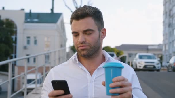帅哥使用智能手机和喝美味的咖啡，而走在办公楼附近的停车场。看起来满意。商业生活方式. — 图库视频影像