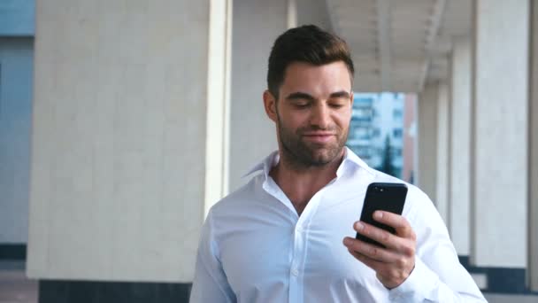 Jonge succesvolle zakenman met behulp van smartphone in de buurt van kantoorgebouw. Bearded knappe man draagt wit shirt. Zakelijke levensstijl. — Stockvideo