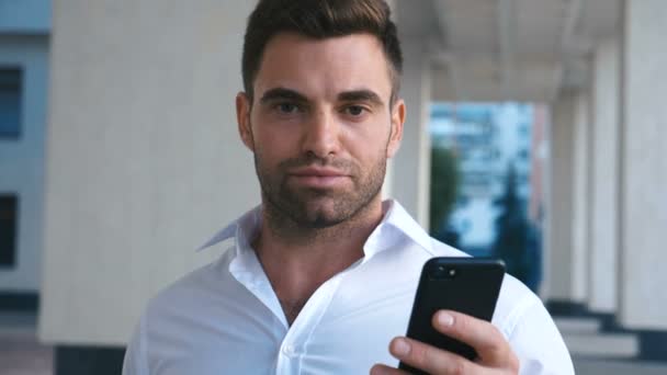 Νέος ελκυστικός επιχειρηματίας χρησιμοποιώντας smartphone κοντά στο κτίριο του γραφείου. Φαίνεται επιτυχημένο. Με γένια όμορφος άντρας φορώντας λευκό πουκάμισο. Επιχειρηματικός τρόπος ζωής. — Αρχείο Βίντεο