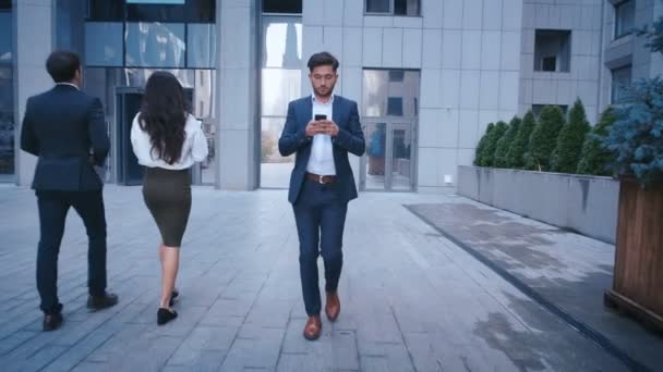 Young Business man använder smartphone medan du går på Big City Business District Street. Klassiskt klädd. Stor kontorsbyggnad i bakgrunden. Ser framgångsrik, säker. — Stockvideo