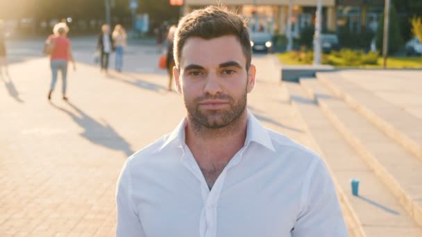 Retrato de un hombre joven y atractivo, con camisa blanca casual, el hombre sonríe y mira a la cámara en el fondo urbano de la ciudad al atardecer. Primer plano de joven hombre de negocios guapo de 30 años al aire libre . — Vídeos de Stock