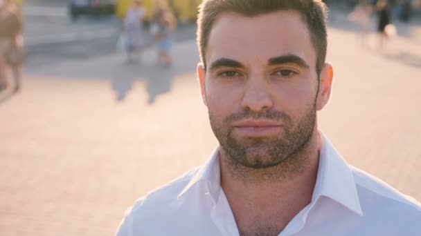 Retrato de un hombre joven y atractivo, con camisa blanca casual, el hombre sonríe y mira a la cámara en el fondo urbano de la ciudad al atardecer. Primer plano de joven hombre de negocios guapo de 30 años al aire libre . — Vídeos de Stock