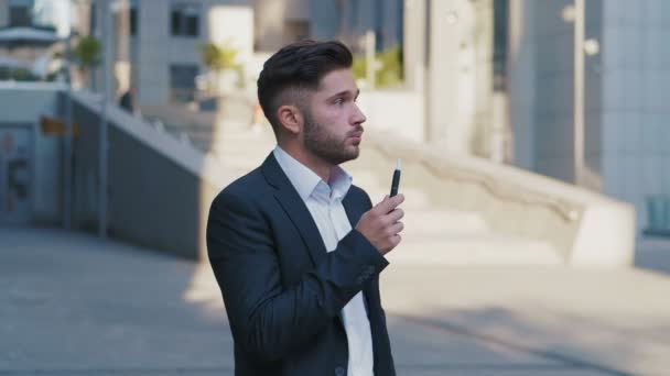 Молодой привлекательный бизнесмен в костюме с помощью смартфона и дымящегося IQOS возле большого офисного здания. Деловая ложь . — стоковое видео