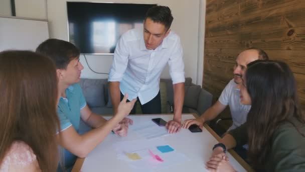 ビジネス チームは、大規模なオフィス テーブルでプロジェクトについて話し合っています。男性チームリーダーがテーブルの近くに立ち、若いクリエイティブチームに指示を与える. — ストック動画